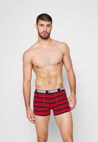 Bañador de boxeo a rayas rojas y negras con un grueso cinturón negro que presenta el logo de Puma. La tela parece suave y elástica.