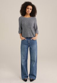 Gråstickad tröja med trekvartslånga ärmar, kombinerad med höga, vida blå jeans och svarta spetsiga klackskor.