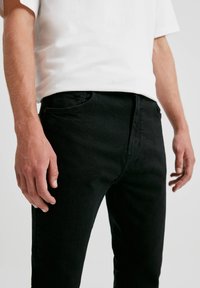 Jeans de mezclilla negra con un corte slim, con bolsillos frontales y detalles de costura sutiles. El material parece texturizado y duradero.