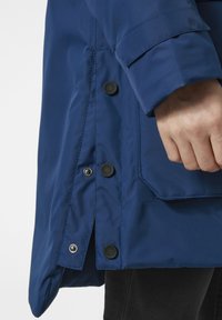Chaqueta azul impermeable que presenta una textura suave, botones con un acabado mate y un bolsillo lateral, con un corte relajado en el dobladillo.