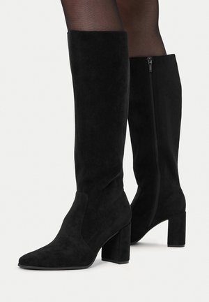 Czarne zamszowe kozaki za kolano na grubych obcasach, noszone z przezroczystymi czarnymi rajstopami, z widocznym zapięciem na suwak z boku jednego buta.