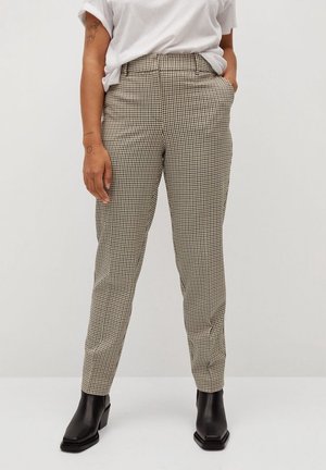 Personne portant un pantalon à carreaux beige et noir, un t-shirt blanc, des bottines noires, debout avec une main dans la poche sur fond blanc.