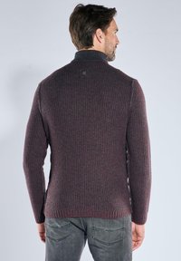 Mann in einem dunkelkastanienbraunen gerippten Pullover mit hohem Kragen, der von hinten schaut, kombiniert mit hellen grauen Jeans vor einem schlichten hellen Hintergrund.