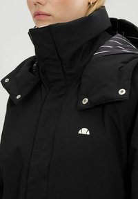 Czarna kurtka z kapturem i wysokim kołnierzem, z zapięciem na napy i białym logo. Wnętrze ma kontrastowy, paskowany wzór.