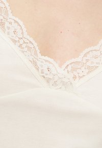 Κοντινό πλάνο ανοιχτόχρωμου δέρματος με λευκό ρούχο διακοσμημένο με φλοράλ δαντέλα κατά μήκος του V-σχήματος λαιμόκοψης.