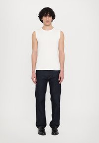 Weißes, geripptes, ärmelloses Oberteil kombiniert mit dunkelblauen, geraden Jeans und schwarzen Schuhen. Der Stoff wirkt glatt mit sichtbarer Textur.