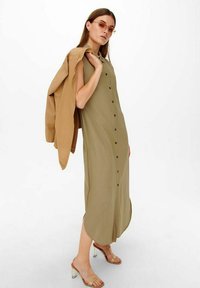 Khaki knoopjurk met een lichte A-lijn vorm, korte mouwen en opgerolde mouwen. Het model draagt een beige jas en doorzichtige hakken sandalen.