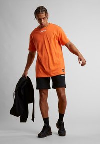 Oranžové bavlnené tričko s voľným strihom, so znakem na hrudi. Model má na sebe čierne šortky a drží čiernu bundu.