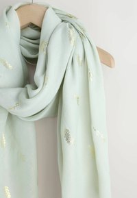 Bufanda verde claro con bordado de hojas metálicas doradas, hecha de tela suave y ligera. Colgada con gracia sobre una percha de madera.