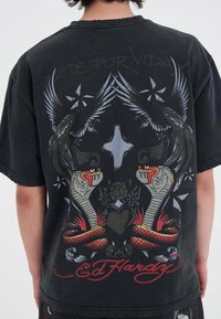Sort t-shirt med et farverigt rygmotiv af slanger, fugle og blomster. Teksten lyder "Arte Por Vida!" med "Ed Hardy" nederst.