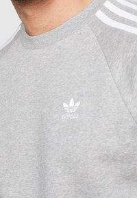 Närbild av en person som bär en ljusgrå sweatshirt med vita ränder på axlarna och en vit broderad logotyp på bröstet.