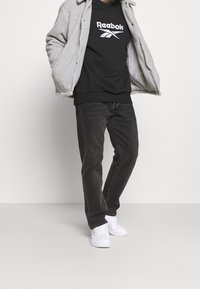 Man i grå fleecejacka, svart Reebok sweatshirt, svarta jeans och vita sneakers som står mot en helt vit bakgrund.