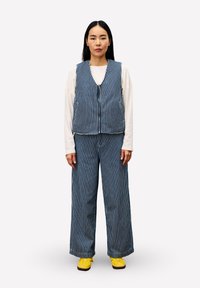 Vrouw staat, gekleed in een blauw-witte verticaal gestreepte vest en broek, een witte blouse met lange mouwen, en gele schoenen tegen een effen achtergrond.