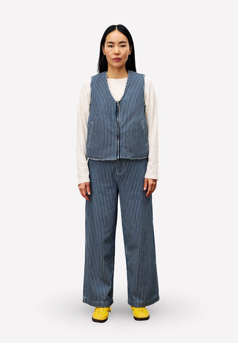 Vrouw staat, gekleed in een blauw-witte verticaal gestreepte vest en broek, een witte blouse met lange mouwen, en gele schoenen tegen een effen achtergrond.