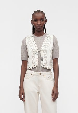 Kvinde med fletninger bærer en hvid broderet vest over en kortærmet striktrøje og højtsiddende hvide bukser, stående mod en ensfarvet baggrund.