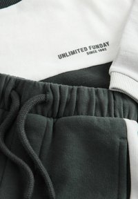 Primo piano di abbigliamento in tessuto bianco e verde scuro con elastico in vita e testo stampato "UNLIMITED FUNDAY SINCE 1982" su fondo bianco.