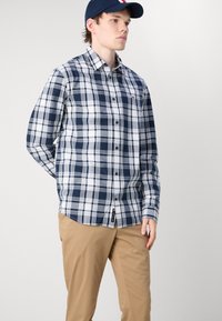 Camisa de cuadros en azul marino y blanco con mangas largas, combinada con pantalones marrón claro. Tela texturizada con un pequeño logo en el pecho.