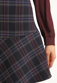 Rutig kjol i marinblått med röda och beige ränder, tillverkad av ett texturerat tyg. Designen inkluderar en åtsittande midja och utsvängd fåll.