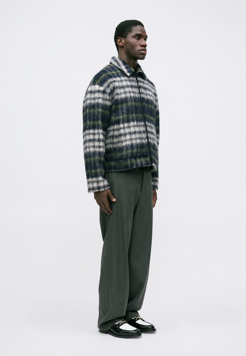 Hombre vestido con una chaqueta de cuadros de felpa con cremallera, pantalones grises holgados y mocasines en blanco y negro, de pie contra un fondo blanco liso.