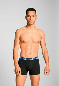 Black boxer briefs with a blue Puma logo waistband. Fabric appears smooth, with a fitted design. Model is shirtless, facing forward.