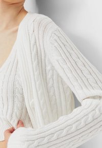 Biały sweter w splot kablowy z dekoltem w serek, prążkowaną fakturą i długimi rękawami, prezentujący skomplikowane wzory oraz miękki materiał.