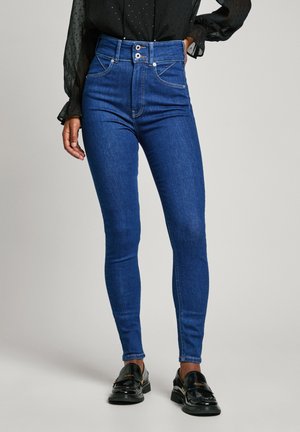 Schlanke, hochtaillierte blaue Jeans mit doppeltem Knopfverschluss, getragen mit schwarzen Loafern und durchsichtigem, langärmligem schwarzem Blusentop.