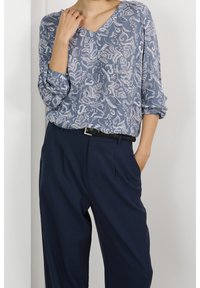 Blouse à motif floral bleu avec un col en V et des manches trois-quarts, associée à un pantalon de costume marine et une ceinture noire. Texture lisse.