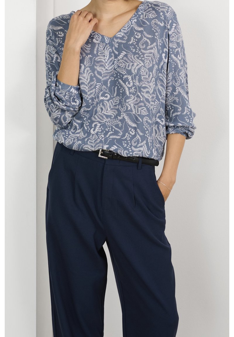 Blouse à motif floral bleu avec un col en V et des manches trois-quarts, associée à un pantalon de costume marine et une ceinture noire. Texture lisse.