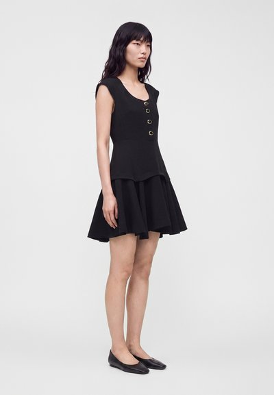 Femme debout portant une robe noire sans manches avec des boutons dorés et une jupe évasée, associée à des chaussures plates noires.