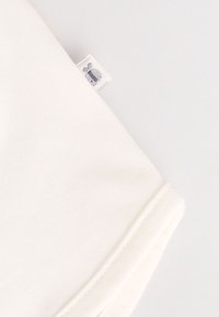 Top de algodón blanco con textura suave, con una pequeña etiqueta gris con un diseño. El dobladillo de la manga está cuidadosamente cosido.