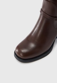 Καφέ δερμάτινη μπότα αστραγάλου με μαύρη σόλα και στρογγυλεμένη μύτη, με οριζόντιο λουράκι κοντά στον αστράγαλο, σε λευκό φόντο.