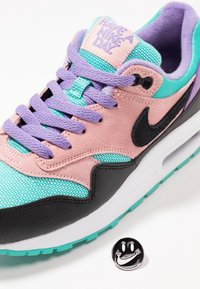 Tenisky Nike Air Max s vrchní částí z růžové semišové kůže a tyrkysové síťoviny, černými akcenty, fialovými tkaničkami a bílou podrážkou. Součástí je pin s usměvavou tváří.