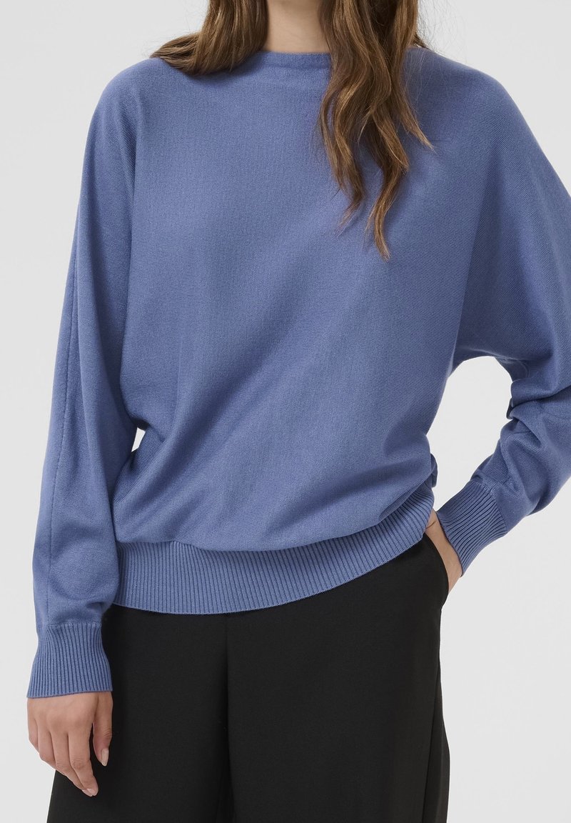 Femme portant un pull bleu à manches longues et un pantalon noir, avec une main dans la poche sur un fond uni.