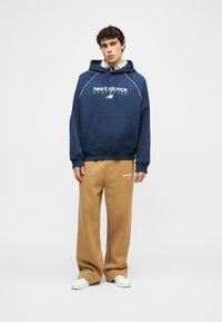 Jonge man die een marineblauwe New Balance-hoodie draagt met beige wijde New Balance joggingbroek en witte sneakers, staand tegen een effen achtergrond.