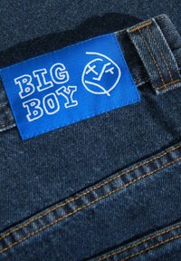 Tejido de mezclilla con una etiqueta azul que dice "BIG BOY" en letras blancas. Presenta costuras naranjas y un corte estándar de jeans. Vista en primer plano.