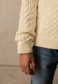 Cremfarbener Strickpullover mit Kabelmuster und gerippten Manschetten, der ein strukturiertes Design aufweist. Getragen über blauen Denim-Jeans, mit einer Hand, die neben dem Körper ruht.