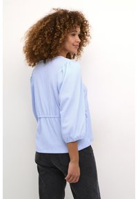 Ljusblå blus med puffärm, texturerad tyg, samlad midja och rund halsringning, i kombination med mörka jeans.
