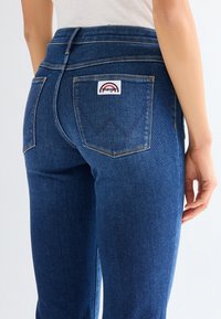 Detail osoby nosící modré džíny Wrangler ukazující zadní kapsy a značkovou nášivku na pravé kapse.