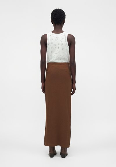 Άτομο που φοράει λευκό αμάνικο τοπ με μαύρα σχέδια και μακριά καφέ φούστα, στέκεται με την πλάτη προς ένα μονόχρωμο φόντο.