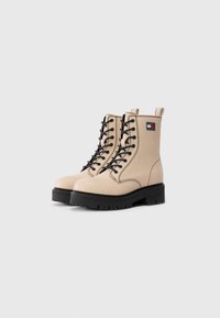 Bottes de combat beige à lacets avec semelles et coutures noires, arborant un patch logo Tommy Jeans sur le côté.