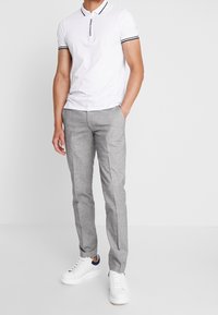 Muž v bílém polo tričku s černým lemem, šedých úzkých kalhotách a bílých teniskách s černým detailem na patě, stojí s rukama v kapsách.