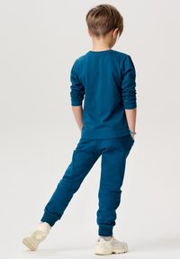 Türkise Langarmshirt und passende Jogginghose aus weichem Stoff. Das Outfit verfügt über enganliegende Bündchen, Taschen und wird mit weißen Sneakern kombiniert.