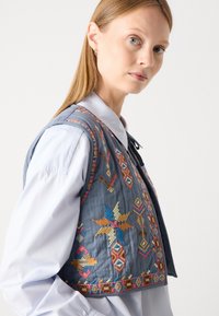 Gilet ricamato blu con motivi geometrici colorati e dettagli, realizzato in tessuto morbido con una finitura testurizzata. Si chiude con un laccio.