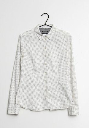 Witte blouse met klein zwart stippenpatroon hangt aan een zwarte hanger tegen een witte achtergrond.