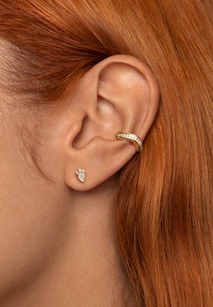 Goldene Creolen mit klaren Edelsteinen, neben einem tropfenförmigen Stecker, der einen weißen Stein und drei kleine Steine darüber hat, getragen von einem Modell mit rotem Haar.