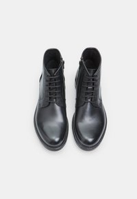 Botas de tobillo de cuero negro con textura suave, diseño con cordones, cremallera lateral y punta redondeada. Presentan costuras mínimas y una suela resistente.