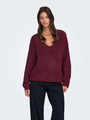 Donna in piedi che indossa un maglione bordeaux a V con trama e pantaloni neri, con capelli castani lunghi ed espressione neutra su uno sfondo semplice.