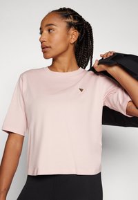 T-shirt curta de manga curta cor-de-rosa claro com decote redondo, apresentando um pequeno logótipo triangular, combinada com leggings pretas e uma mala de ombro.