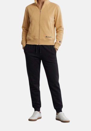 Personne portant une veste beige zippée avec le logo Champion, un pantalon de jogging noir et des baskets blanches, debout les mains dans les poches.