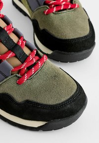 Zielone zamszowe sneakersy z czarnymi akcentami, posiadające grube czerwono-białe sznurówki, teksturowane wykończenie oraz gumową podeszwę o zaokrąglonym kształcie.